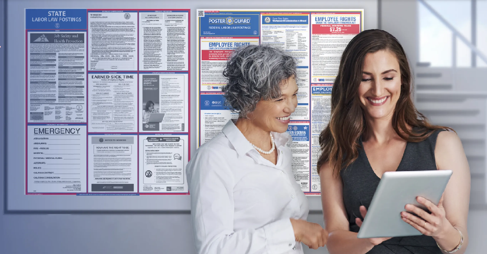 Two women looking at a tablet with a bulletin board of notices in the background